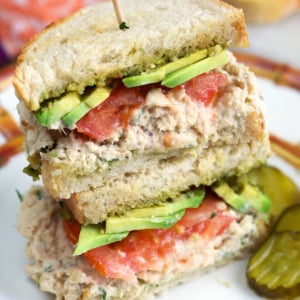 Tunacado sandwich on sourdough bread on a plate with pickles on the side.