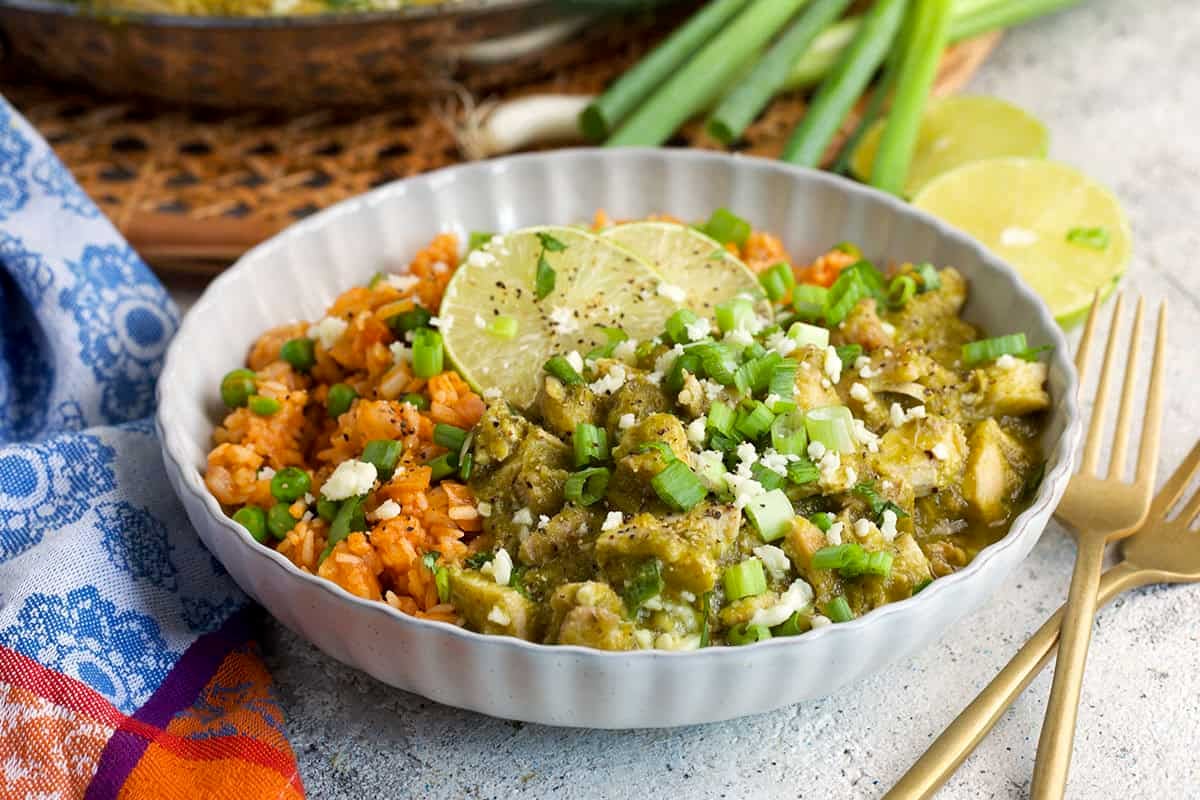 Chicken Chile Verde served in a shallow white bowl over Mexican rice and topped with limes, cilantro and cotija cheese.