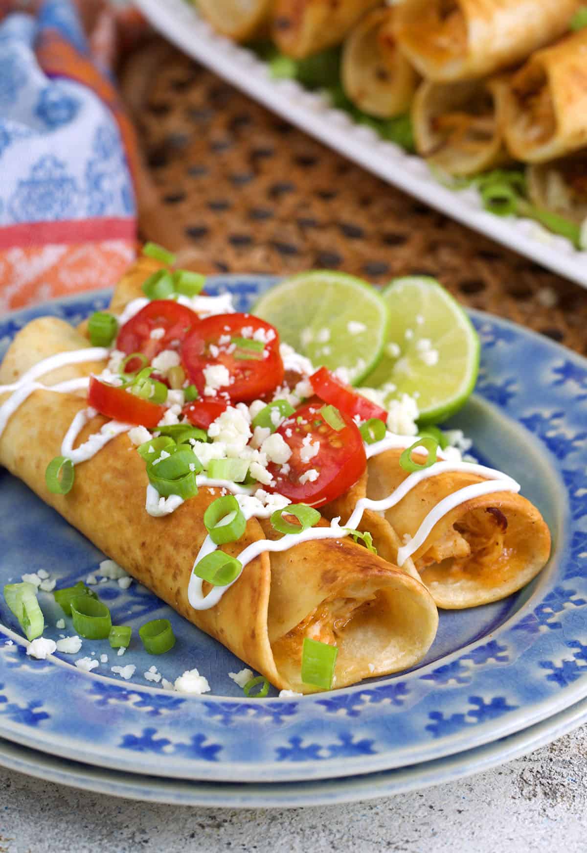 Two chicken flautas on a blue plate topped with sour cream, tomatoes and green onions.