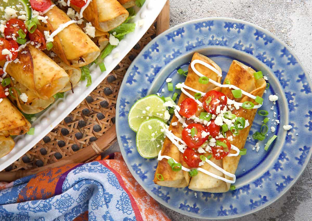 chicken flautas being served on a blue plate with a platter of flautas to the left.