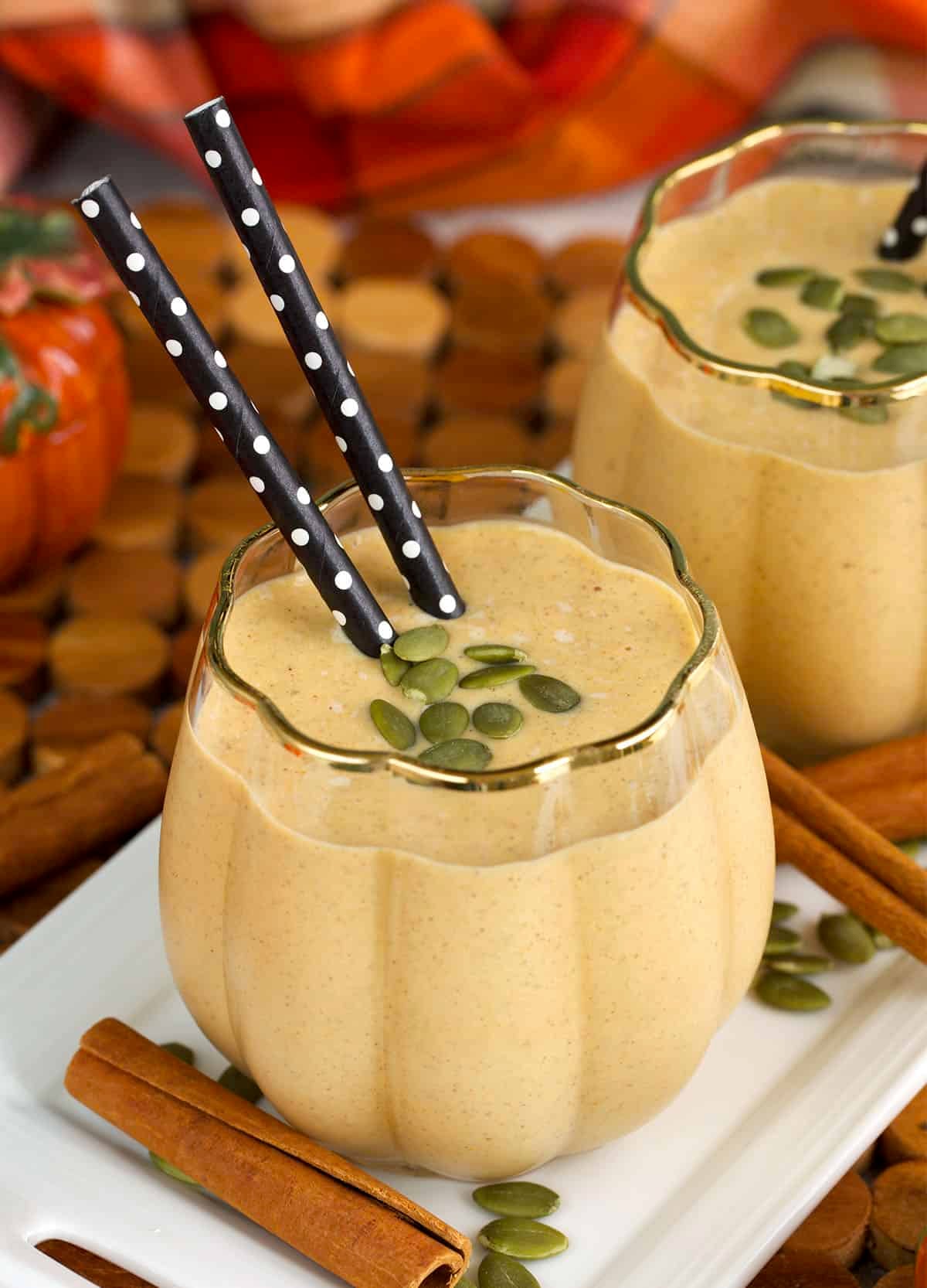 Pumpkin smoothie on a white tray, topped with pumpkin seeds and two straws.