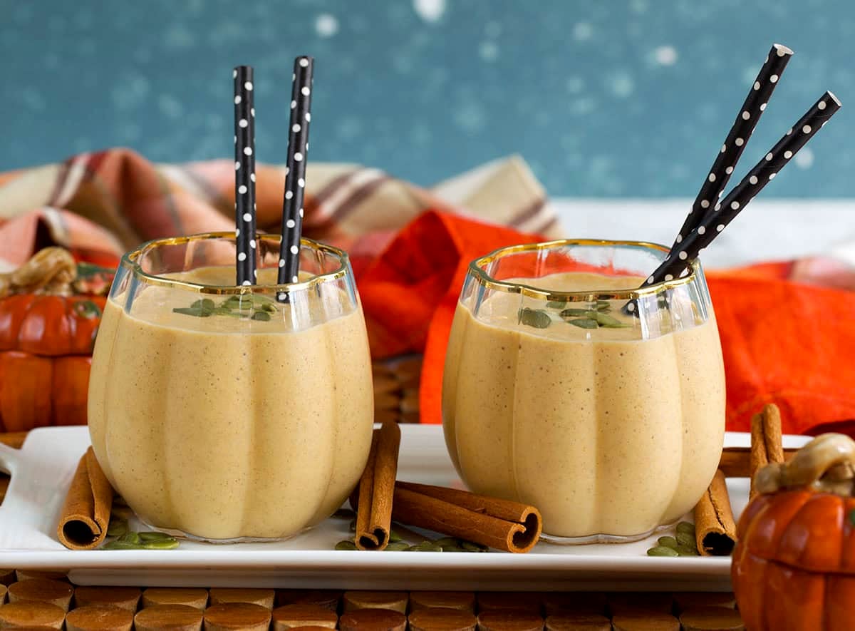 two pumpkin glasses filled with pumpkin smoothies on a white tray with cinnamon sticks and pumpkin seeds.